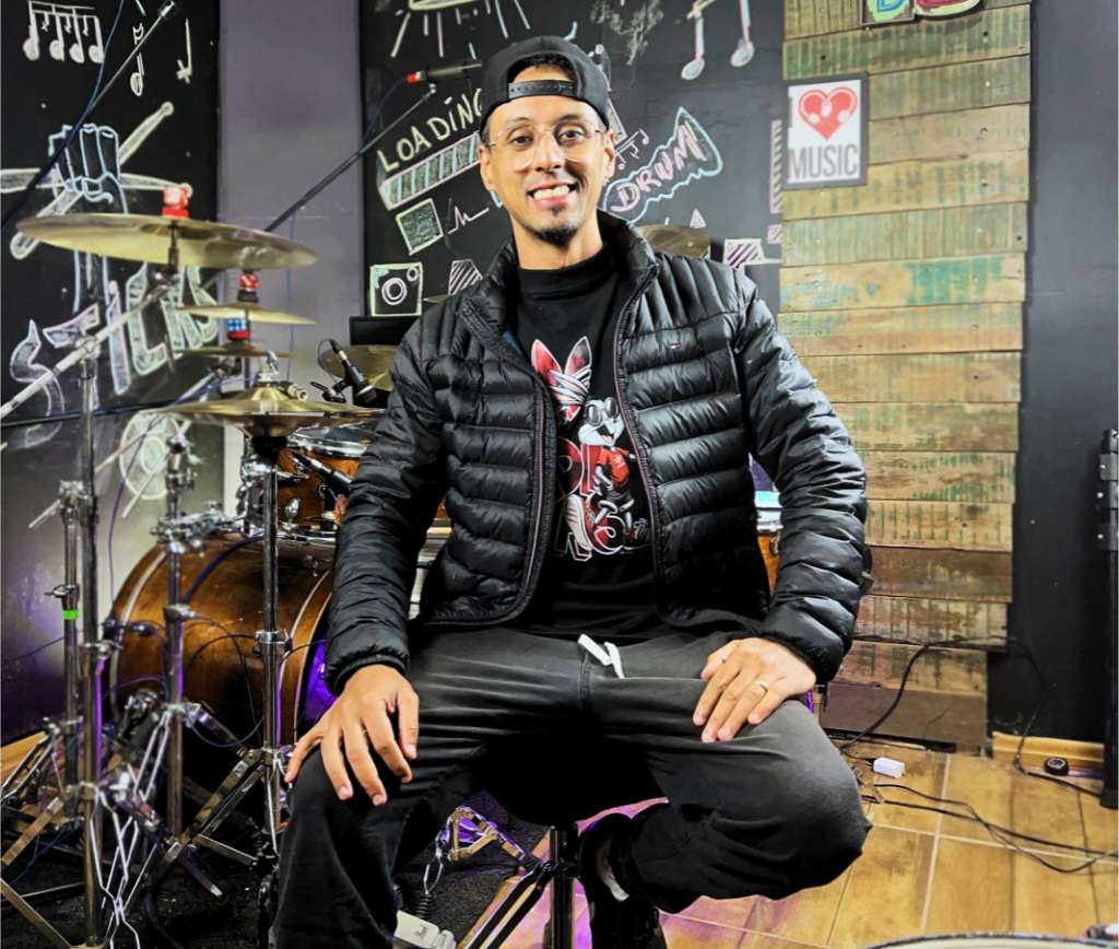 Chikito Borges, baterista e professor, sentado em frente a uma bateria no estúdio